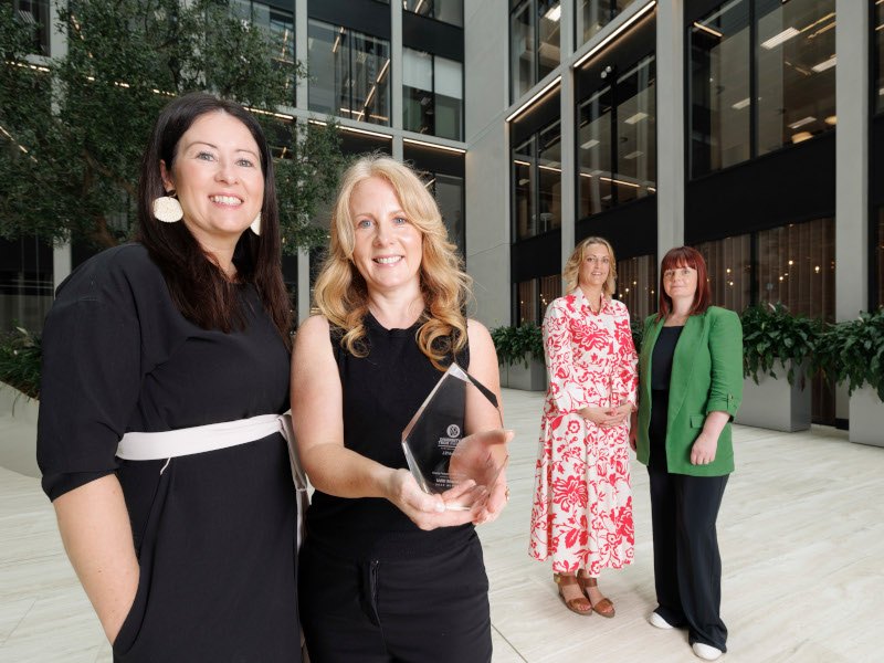 Four women standing in front of buildings and a tree. One of the women is holding an award in her hands.