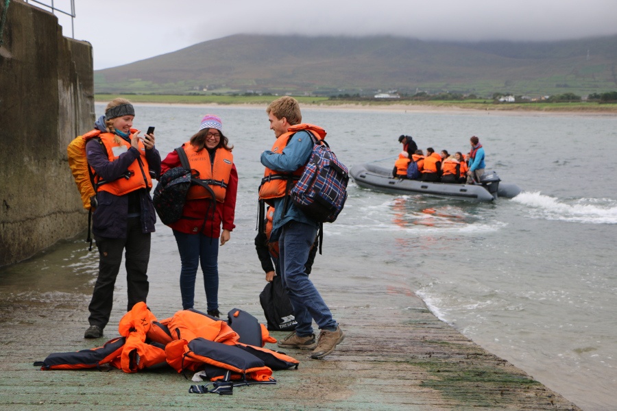 A group of people on a pier wearing orange lifejackets with a small boat behind them in the water.