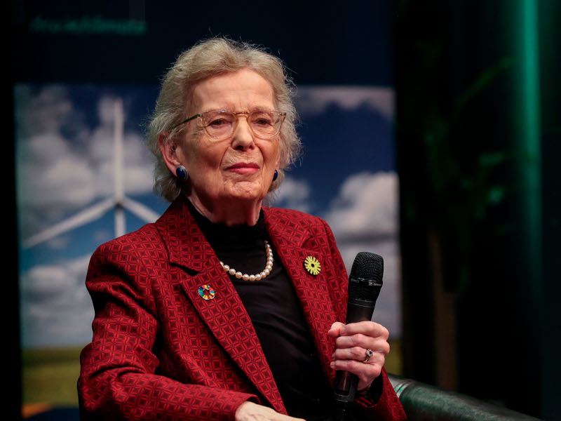 A woman wearing a red jacket and a black shirt looks ahead while holding a microphone in her left hand.