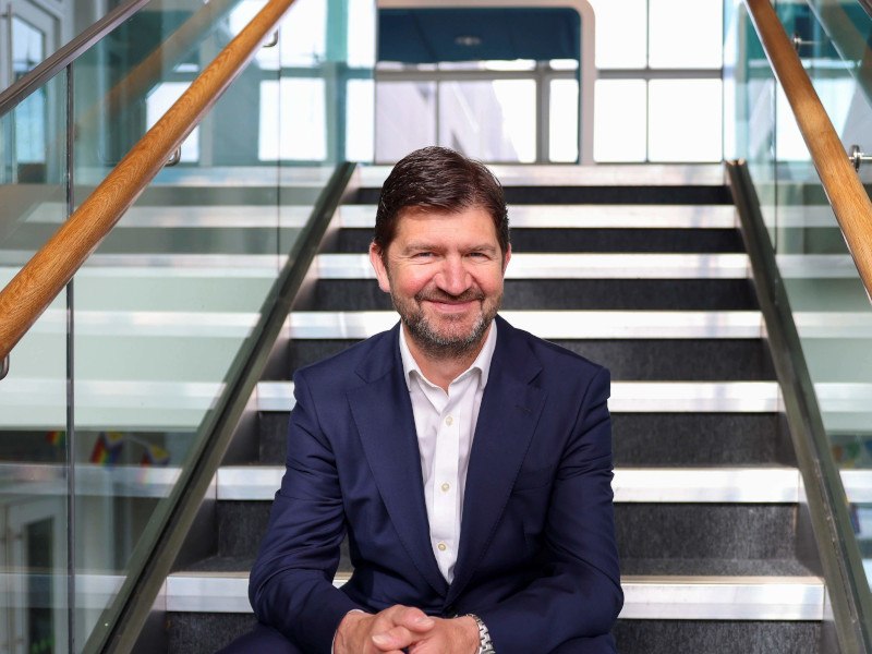 Eir CEO Oliver Loomes wearing a navy suit and sitting on some stairs in a building.