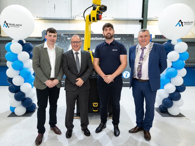 Four men stand smiling in front of an automated machine with pillars of balloons situated at either side of the men.