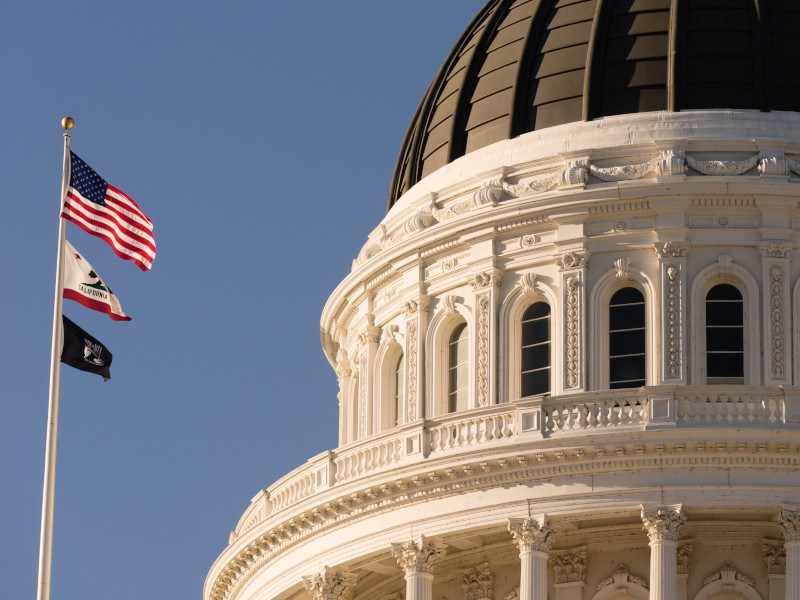 Photo of California State Capital building with a US flag among flags flying next to it.