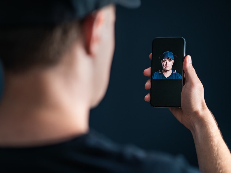 A man holds a phone in his hand with the display showing his face. Represents the process of creating AI deepfakes.