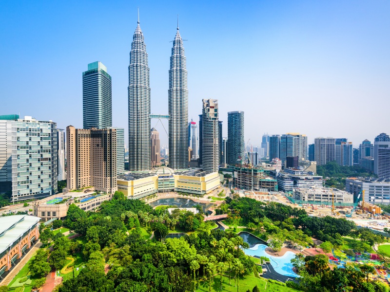 A photo of Kuala Lumpur's skyline with skyscrapers in the background against a bright blue sky and a green park scene in the foreground.