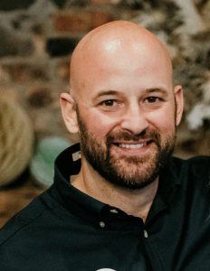 A cropped photo of Raymond Bond smiling at the camera in a black shirt, with a brick wall behind him.