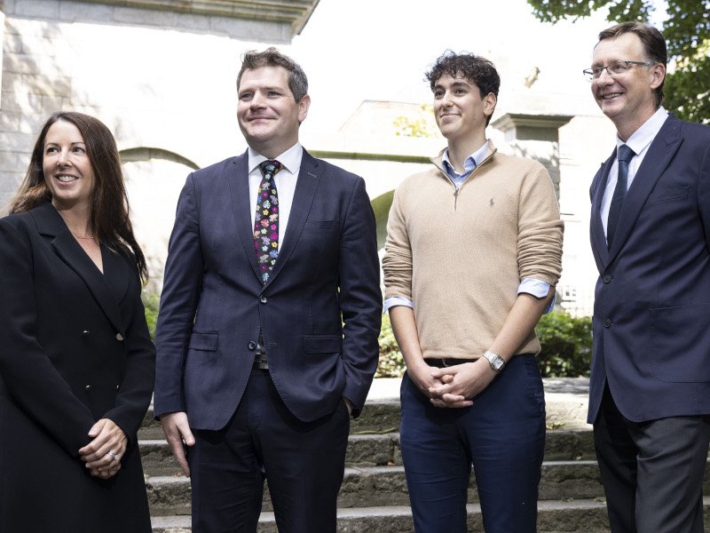 A woman and three men stand in a row and smile towards a camera.