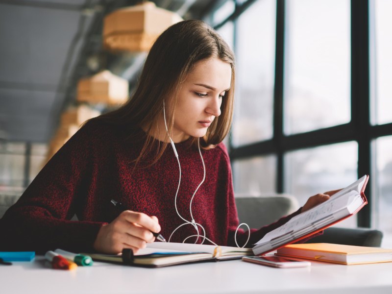 A student sits at a desk in a library reading and taking notes with her headphones in. She looks like she's concentrating.