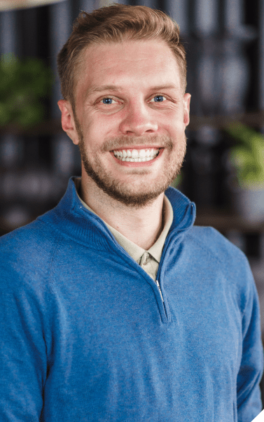 A man wearing a blue jumper smiles at the camera in an office setting.