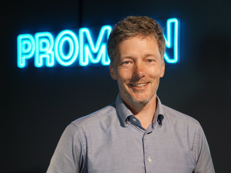 A man wearing a light blue shirt smiles at the camera in front of a blue neon sign displaying the company name, Promon.