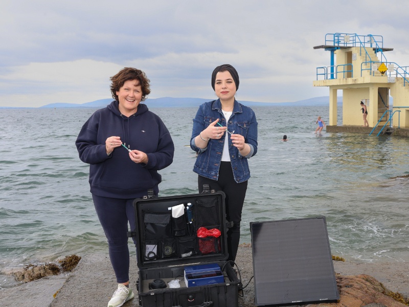 Two women standing in front of the sea holding water testing kits in their hands.