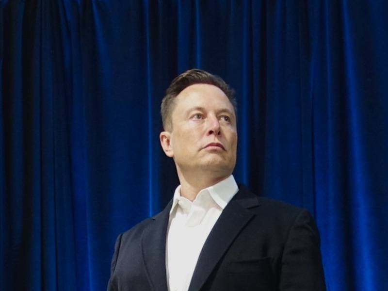 Elon Musk wearing a suit and standing on a stage with a dark blue curtain behind him.