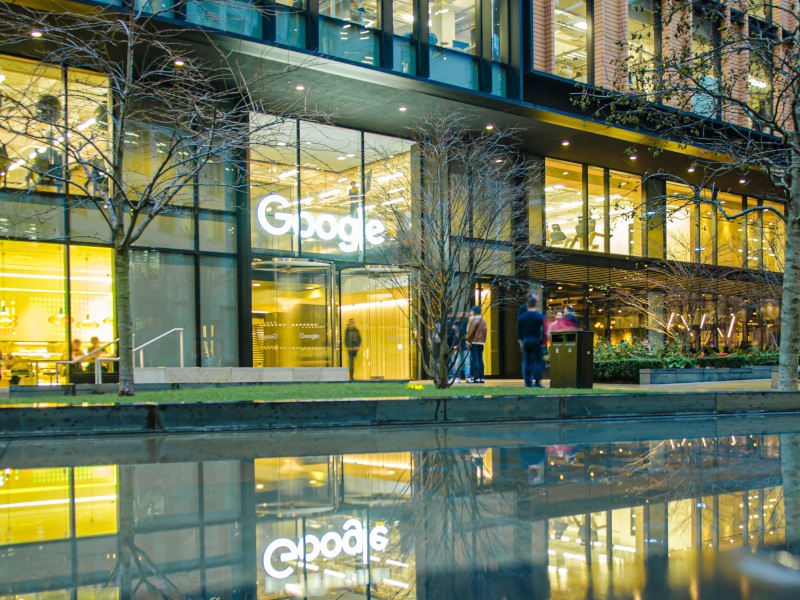 The Google logo on the side of a building, with lights visible through multiple windows and water in front of the building.