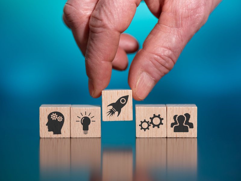 A hand picking out a wooden cube with a rocket sign on it signifying a start-up.