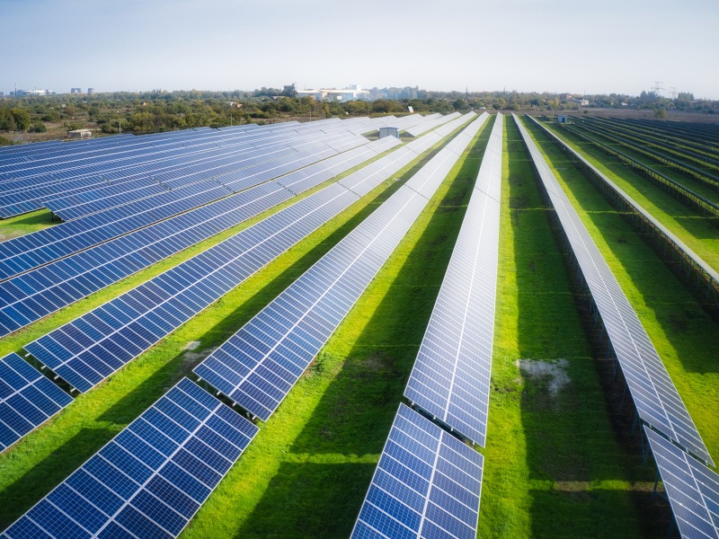 Multiple rows of solar panels on a field with the sun shining above.