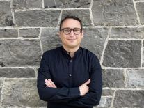 Dr Andreas Balaskas stands in a black shirt with his arms folded against a grey stonewall and smiles at the camera.