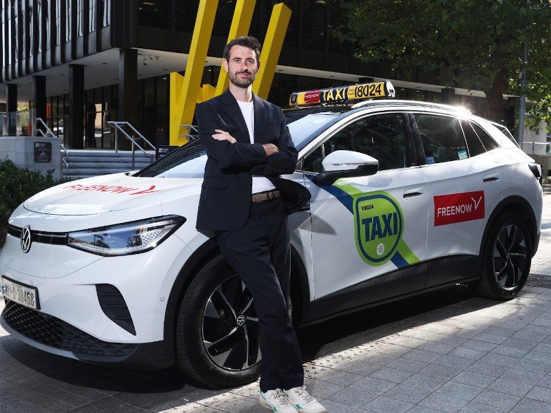 A man wearing a suit, t-shirt and runners stands with arms folded leaning against a white taxi with a Free Now sticker on the back door.