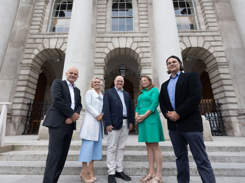 In a low angle shot, five people are standing in front of the Trinity College Dublin building. There are three people in suits and two in dresses in between.