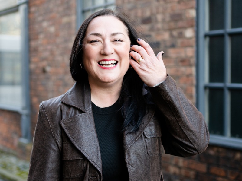 A woman wearing a brown leather jacket, smiling at the camera, while pulling her hair back.