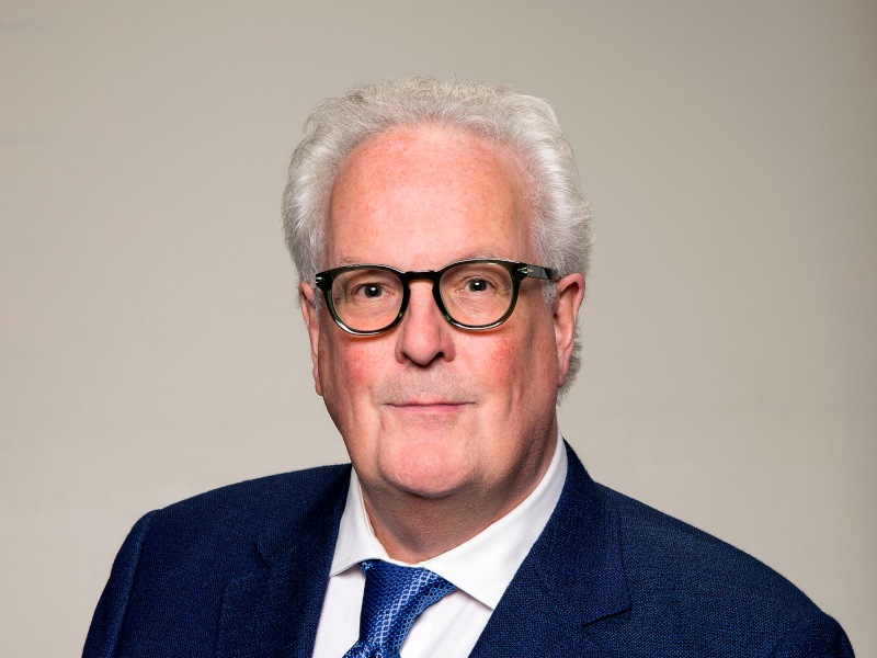 A man wearing a suit and tie with glasses smiles at the camera in front of a grey background.