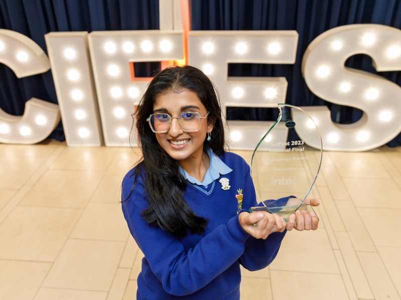 A young female student in a blue school uniform holding a glass award in front of 3D lit-up letters that spell SciFest