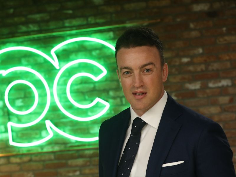 A young man wearing a suit smiles at the camera standing in front of a light sign of green lettering on a brick wall.