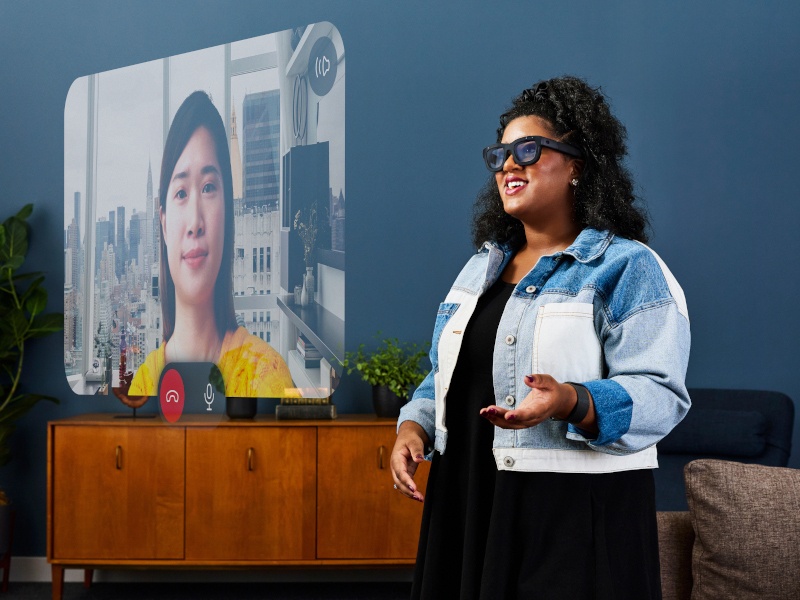 A woman wearing a pair of black AR glasses stands in a room. In front of her is a holographic screen with another person on it.