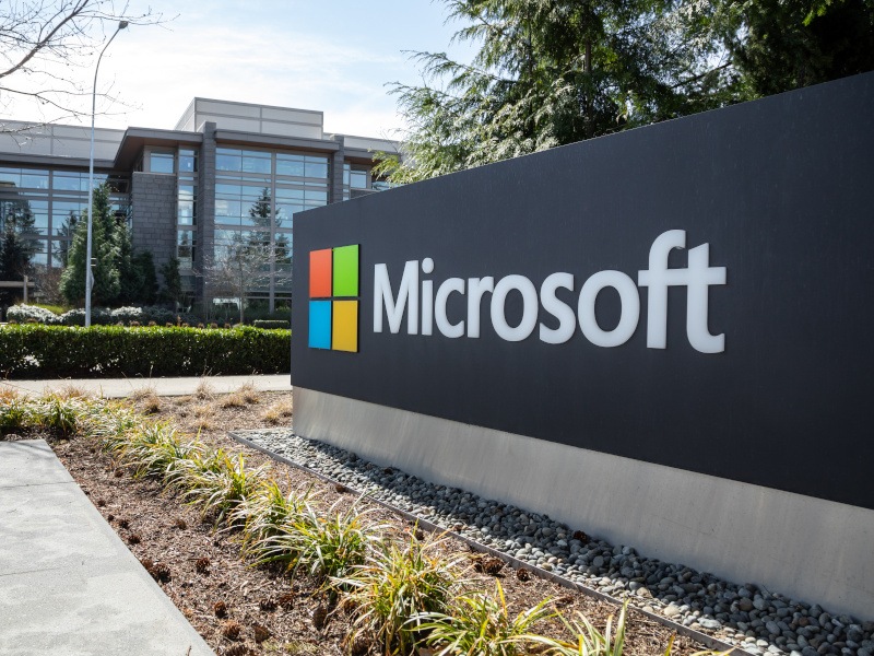 A close-up of a large sign with the Microsoft logo. In the background, there is a large glass office building.