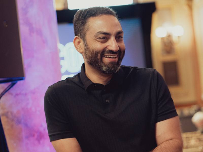 A man wearing a black short-sleeved shirt smiles while looking out of frame. He appears to be at a conference.