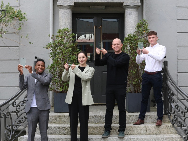 Four people holding awards with both their hands stand diagonally on a small staircase.