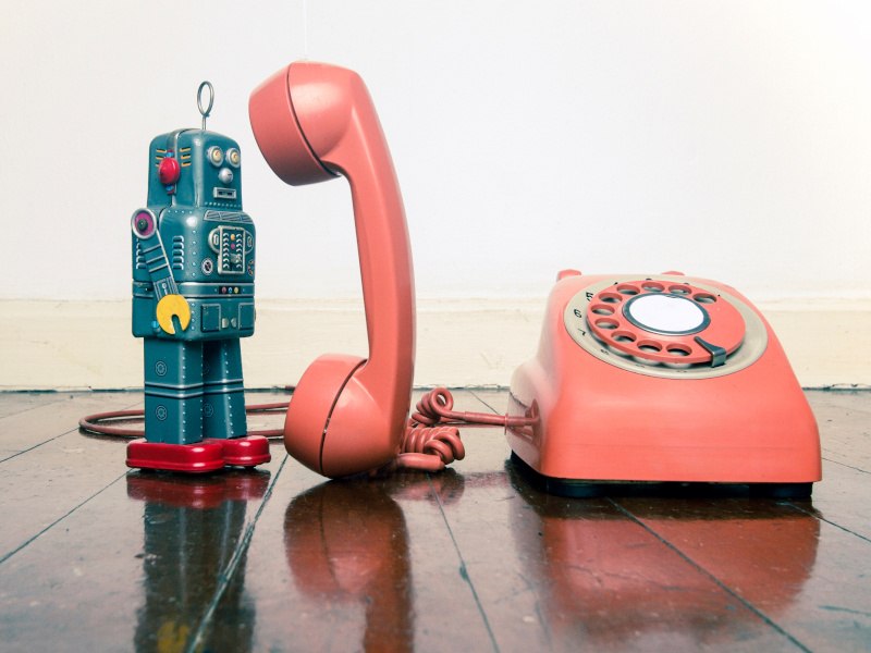 A toy robot stands on a floor beside a vintage pink telephone. The receiver is standing upright facing the robot, symbolising robocalls.