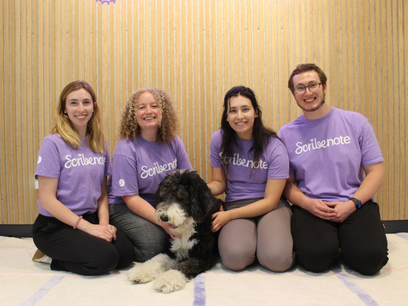 Three women and a man wearing purple t-shirts with the Scribenote logo on it. They sit on the floor with a large black and white dog.