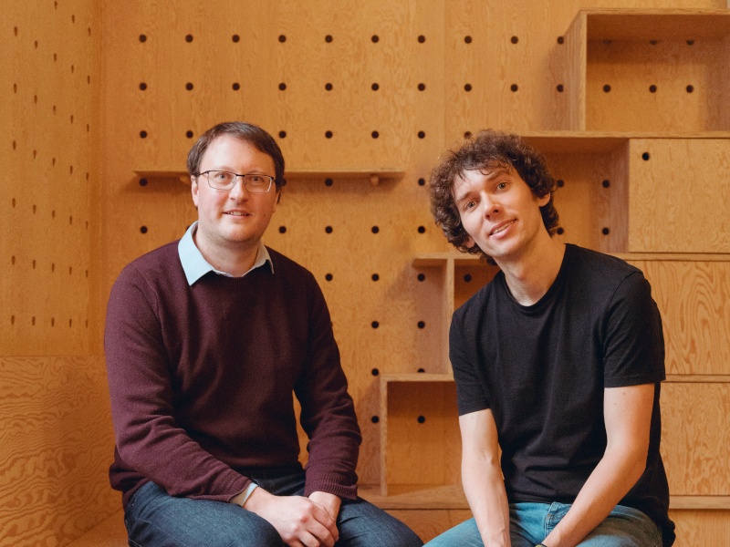 Two men sit side by side in front of an orange-coloured wall adorned with empty shelves.