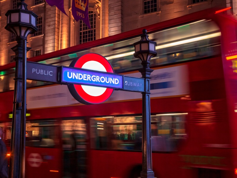 A sign for the London Underground subway between two lampposts, while a blurry red double decker bus whizzes passed on a city road at night.