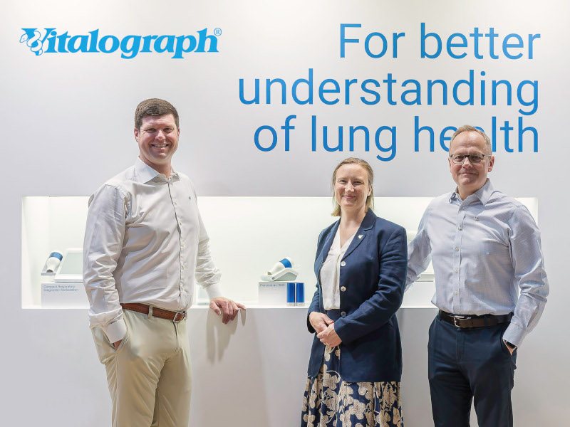 Three people standing in front of a science-lab like backdrop with the 'Vitalograph' logo on the top left corner. One woman is standing in the middle of two men.