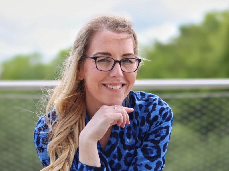 A close-up headshot of Alison O'Connor. She is wearing a blue and black patterned blouse and has long blonde hair and glasses. Her chin is resting on her hand.