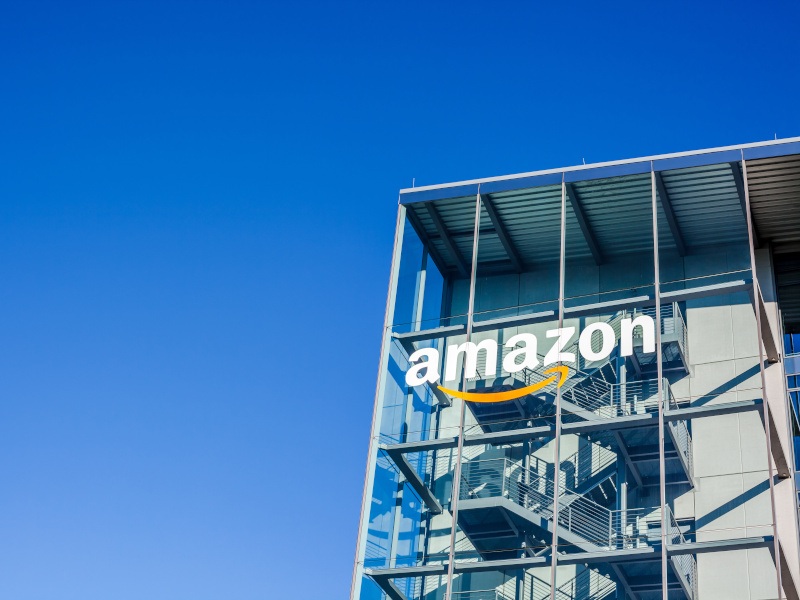 A photo from below of a glass building with the Amazon logo on it, against a clear blue sky.