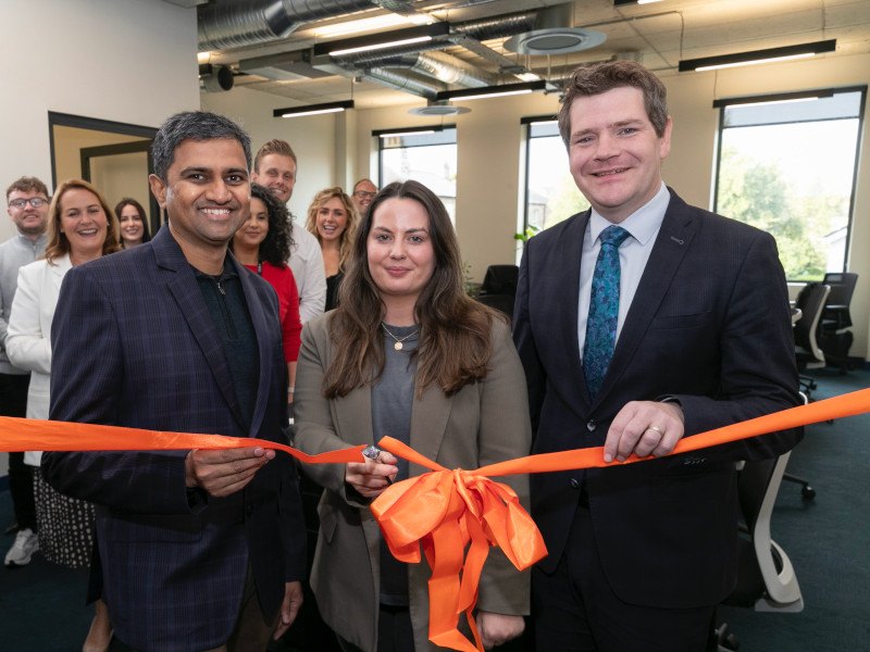 Multiple men and women standing together as an orange ribbon is cut in an office room. The opening of the Chargebee office in Dublin.