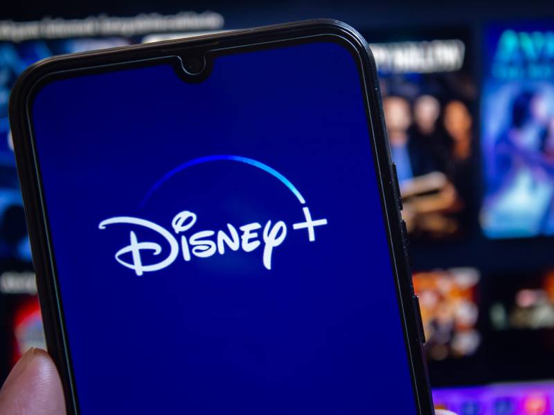 An out of focus Disney+ homepage with a smartphone in the foreground displaying the Disney+ logo.