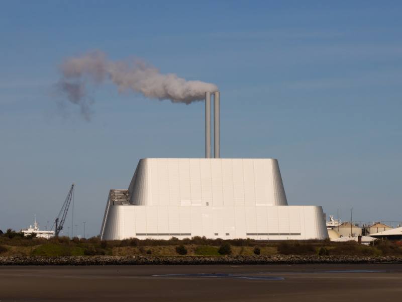 The Poolbeg waste incinerator on a warm day. The building is grey in colour and is releasing a thick dark grey smoke from the chimney on top.