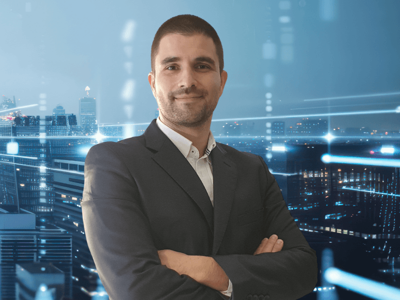 Jose Carlos Pereira stands with his arms folded smiling at the camera against a superimposed cityscape grey-blue background.