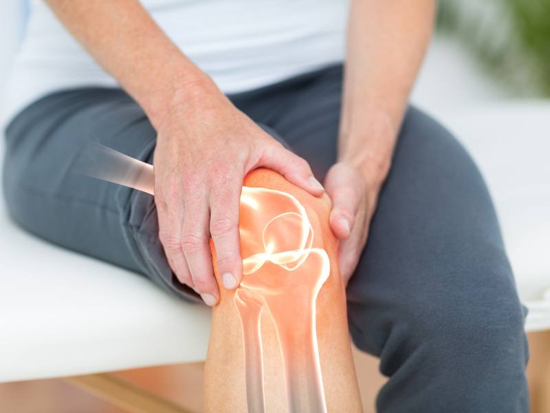 An image of the torso of a man sitting down, holding his knee with an illustration of the knee bones superimposed on the image.