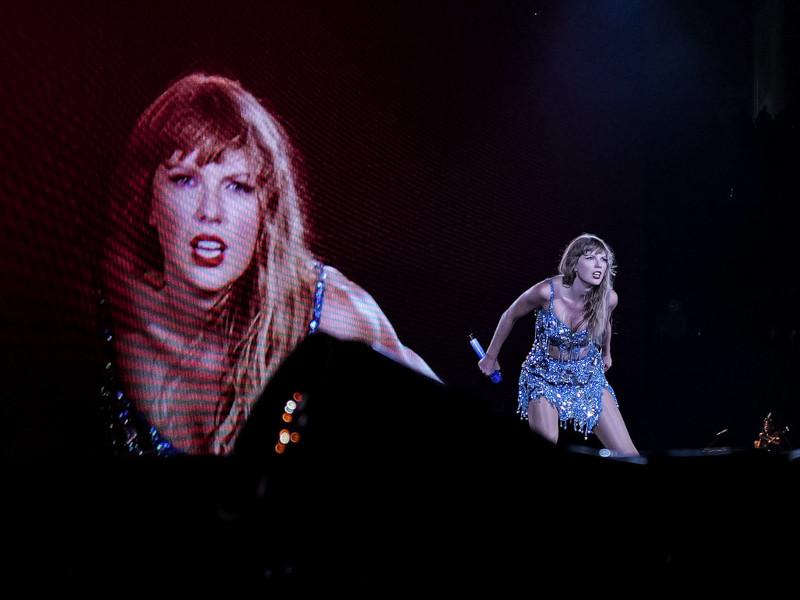 Taylor Swift performs on stage in a blue outfit with a large screen behind her showing a close-up of her face.