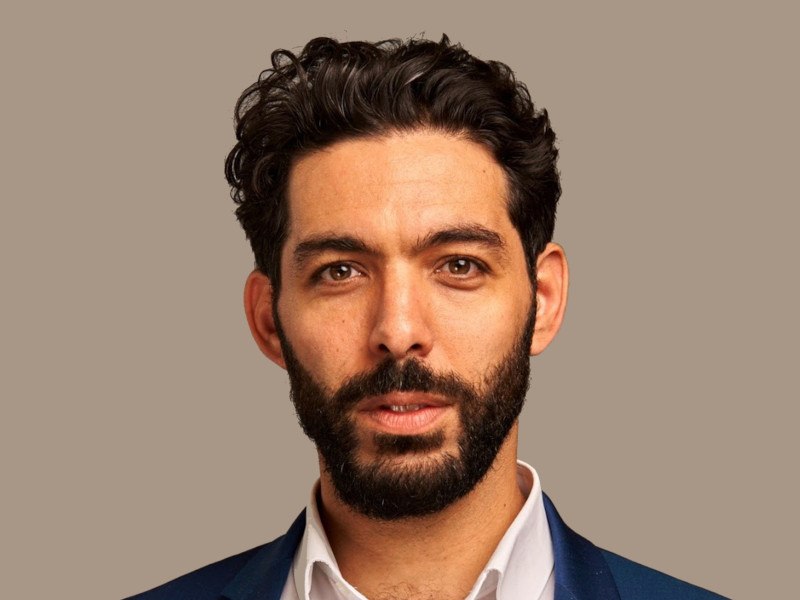 A man with a beard wearing a suit looks at the camera over a beige coloured background.