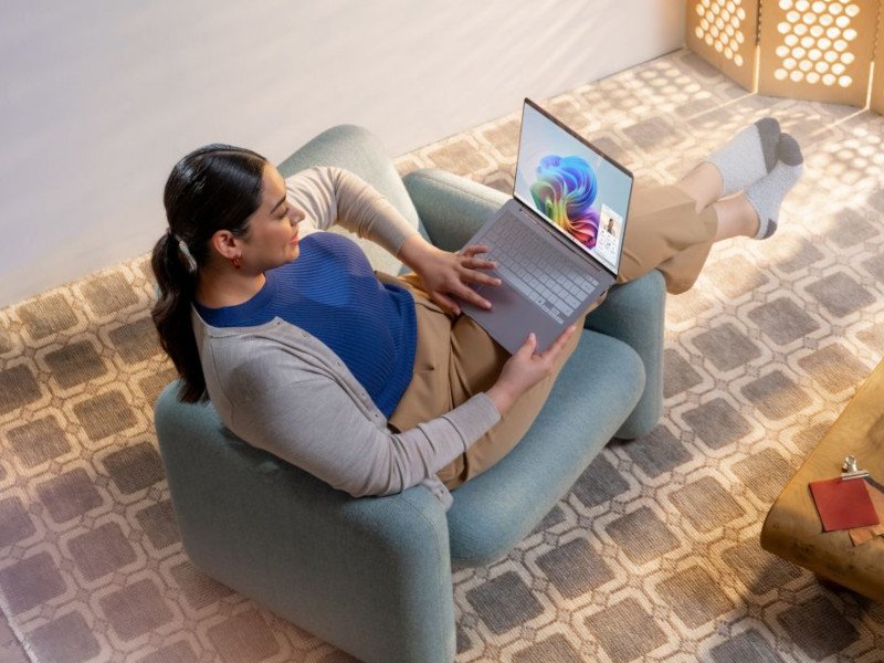 A woman sits with her legs over the arm of an armchair with a laptop on her lap that has Microsoft Copilot open on it.