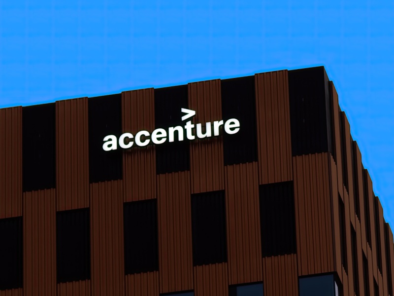 In a high contrast picture, the sky is bright blue and a brown and black building with sharp edges is in the front with the Accenture logo in white on it.