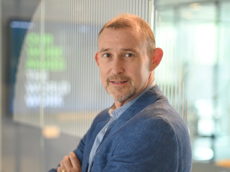 A side-on shot of Adam Spearing, who looks at the camera with his arms folded wearing a light-blue suit jacket in an office.