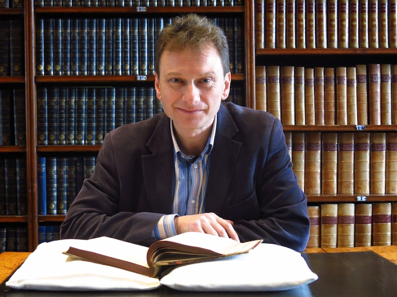 A man sits at a table with an open book in front of him. Behind him are shelves full of books.