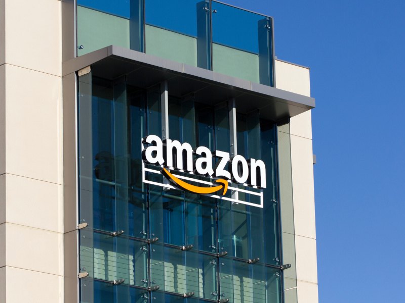 The Amazon logo at the top of a large building with a lot of glass against a bright blue sky.