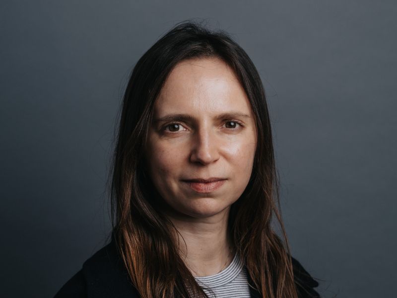 A woman with long brown hair smiles at the camera over a dark grey background.