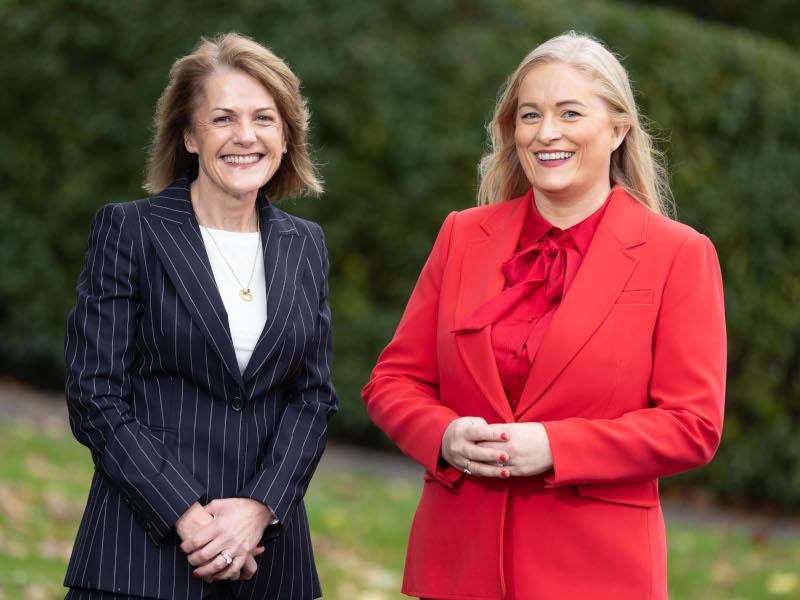 Two women in business suits stand side by side smiling at the camera in an outdoor area.